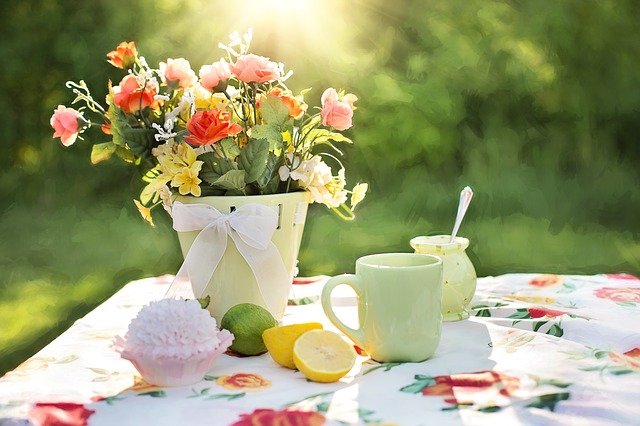 A sunlit outdoor table with a floral tablecloth sets the scene for a summer garden party, topped with a bouquet of pink and yellow flowers in a pale bucket, a green mug, a jar with a spoon, a cupcake, and slices of lemon and lime.