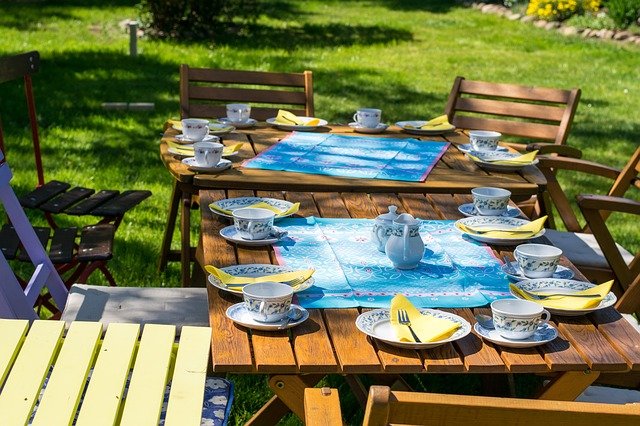 A wooden outdoor table set for tea with cups, saucers, plates, and yellow serviettes on blue place mats—perfect for a summer garden party. Surrounded by chairs in a sunny garden with green grass and flowers in the background.