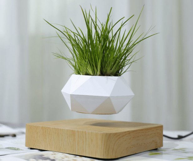 A geometric white planter with green grass floats above a square wooden base, appearing to levitate—a striking piece of levitating décor. The background is softly blurred with light-coloured curtains, perfect for modern interior design.