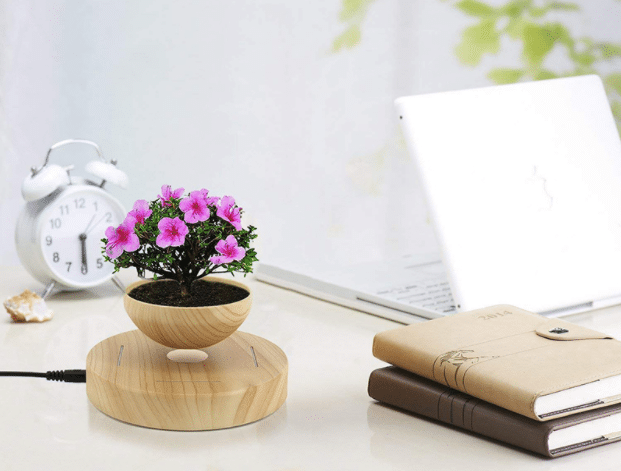 A small bonsai tree with pink flowers levitates in a wooden pot on a white desk, near an alarm clock, closed laptop, and two notebooks—showcasing how levitating décor is shaping the future of interior design in bright, minimalistic spaces.