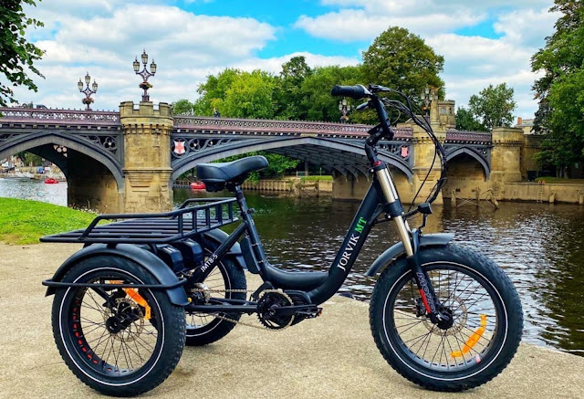 A black Jorvik electric tricycle, one of the leading adult tricycles, is parked on a path beside a river with a decorative stone bridge and trees in the background under a partly cloudy sky.