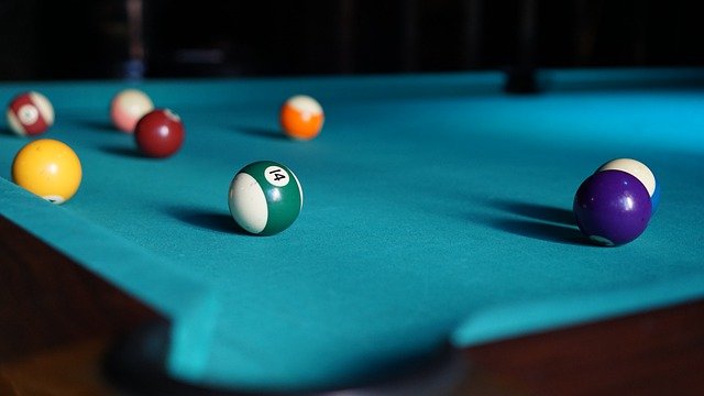 A close-up of a blue pool table shows several billiard balls scattered across its surface, with the green-striped 14 ball prominently in the foreground and other coloured balls dotting the pool table in the background.