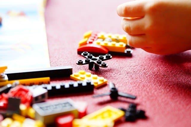 A child's hand reaches towards scattered colourful LEGO pieces on a red surface, with partially assembled bricks and an instruction manual visible—highlighting the role of children's toys in cognitive development.
