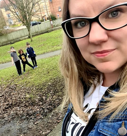A woman wearing glasses takes a selfie outdoors; in the background, three children stand together on grass near a path and some buildings, capturing one of the simple ways to bond with your children.