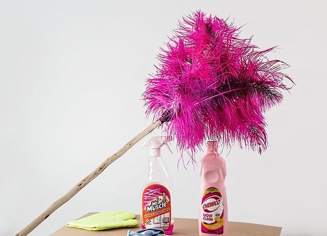 A pink feather duster leans against two cleaning spray bottles on a table, with a yellow cloth nearby—ready for a spring clean. The background is plain and white.