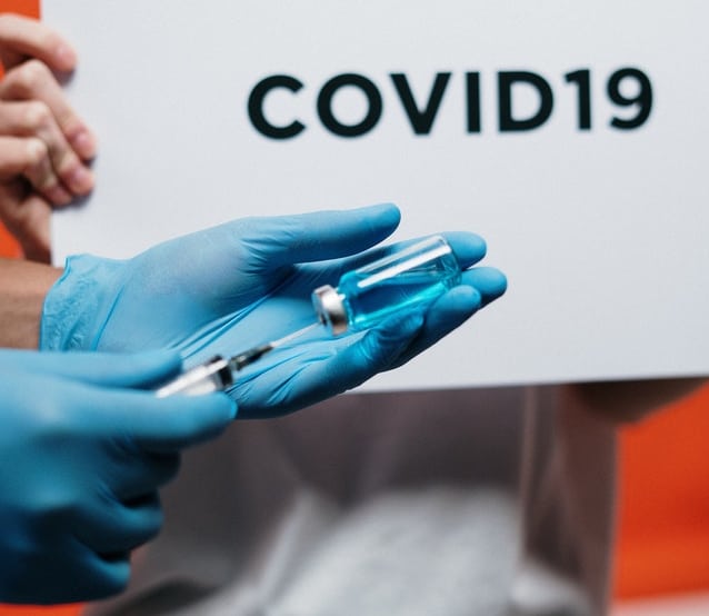 A person wearing blue gloves holds a vial and a syringe filled with blue liquid in front of a sign that reads "COVID19," highlighting the administration of a covid-19 vaccine.