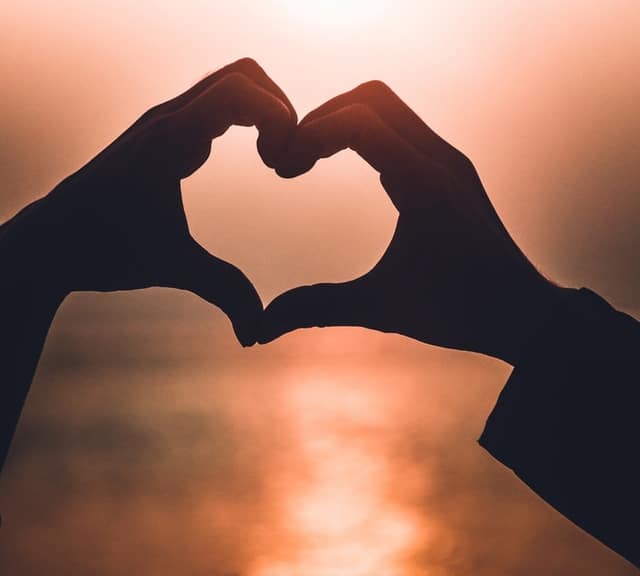 Two hands form the shape of a heart against a soft, warm sunset—an inspiring scene for your next at-home date night idea, with the sun’s glow creating a romantic silhouette effect.