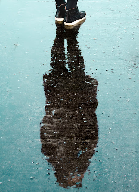 A person stands on wet pavement, visible from the knees down, their reflection clear in a puddle speckled with raindrops—a quiet reminder to stop victim blaming and prioritise women's safety in every environment.