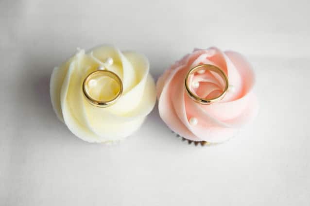 Two cupcakes with rose-shaped icing, one white and one pink, each topped with a gold wedding ring and a small edible pearl—an elegant nod to UK wedding themes and extravagant wedding cakes on a white background.