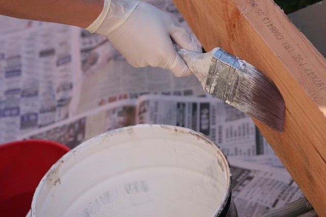 A gloved hand uses a paintbrush to apply white paint to a wooden surface—a simple step in home improvement. An open tin of paint and newspapers used as protective covering are visible in the background.
