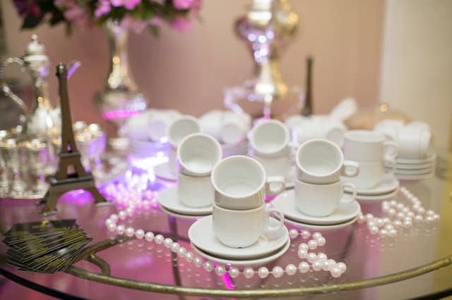 A close-up of a glass table set with stacked white coffee cups and saucers, a string of pearls, small Eiffel Tower figurines, and blurred pink flowers in the background captures an elegant Gatsby Party ambience.