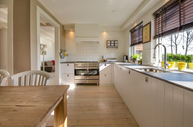 Bright, modern kitchen with white cupboards, a wooden dining table, and large windows with brown blinds letting in natural light. Potted plants and stylish kitchen appliances offer inspiring kitchen ideas to make your kitchen look expensive.