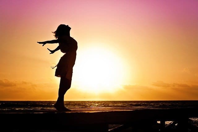 Silhouette of a woman standing on a beach with arms outstretched, facing the sunrise or sunset over the sea, as a bright golden sky glows—symbolising a boost in confidence and self-esteem.