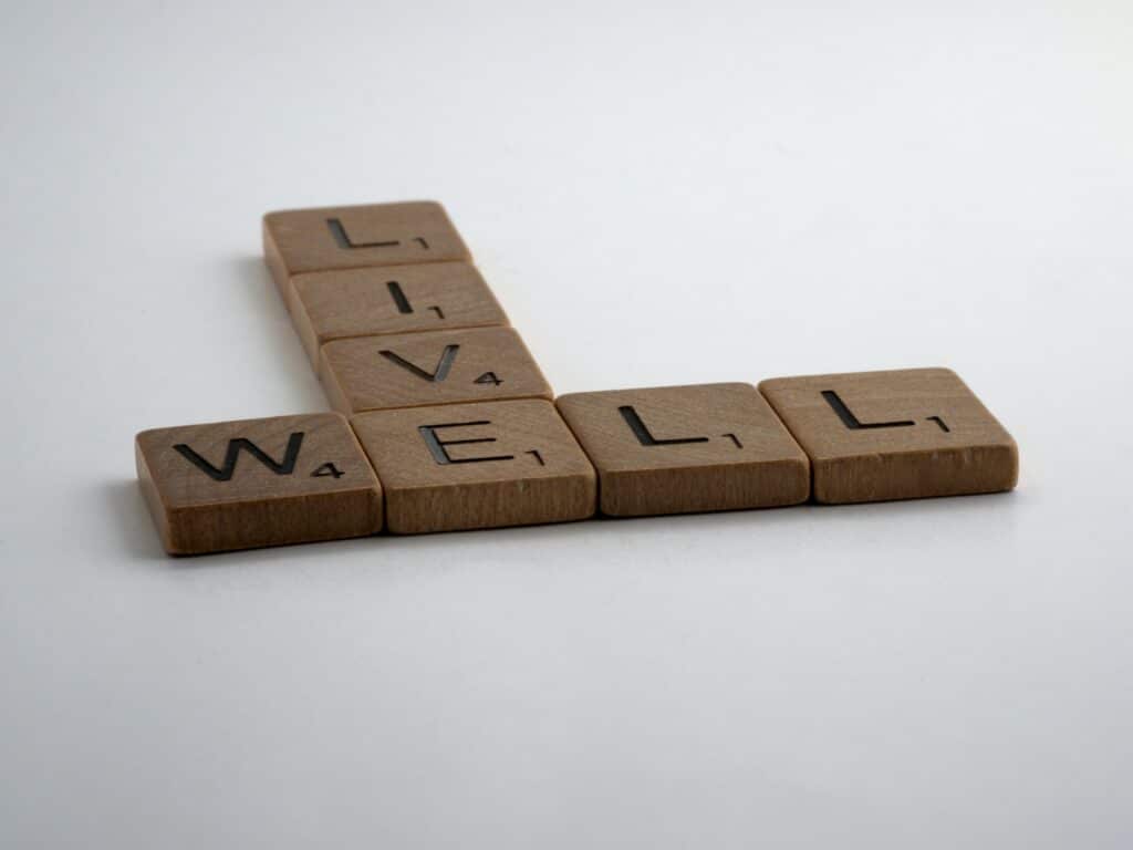Four Scrabble tiles spelling "WELL" are arranged horizontally, with "LIVE" intersecting the "L," symbolising the importance of mental wellbeing. The tiles are set against a plain white background.