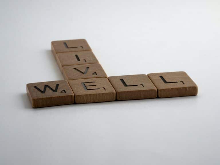 Four Scrabble tiles spelling "WELL" are arranged horizontally, with "LIVE" intersecting the "L," symbolising the importance of mental wellbeing. The tiles are set against a plain white background.