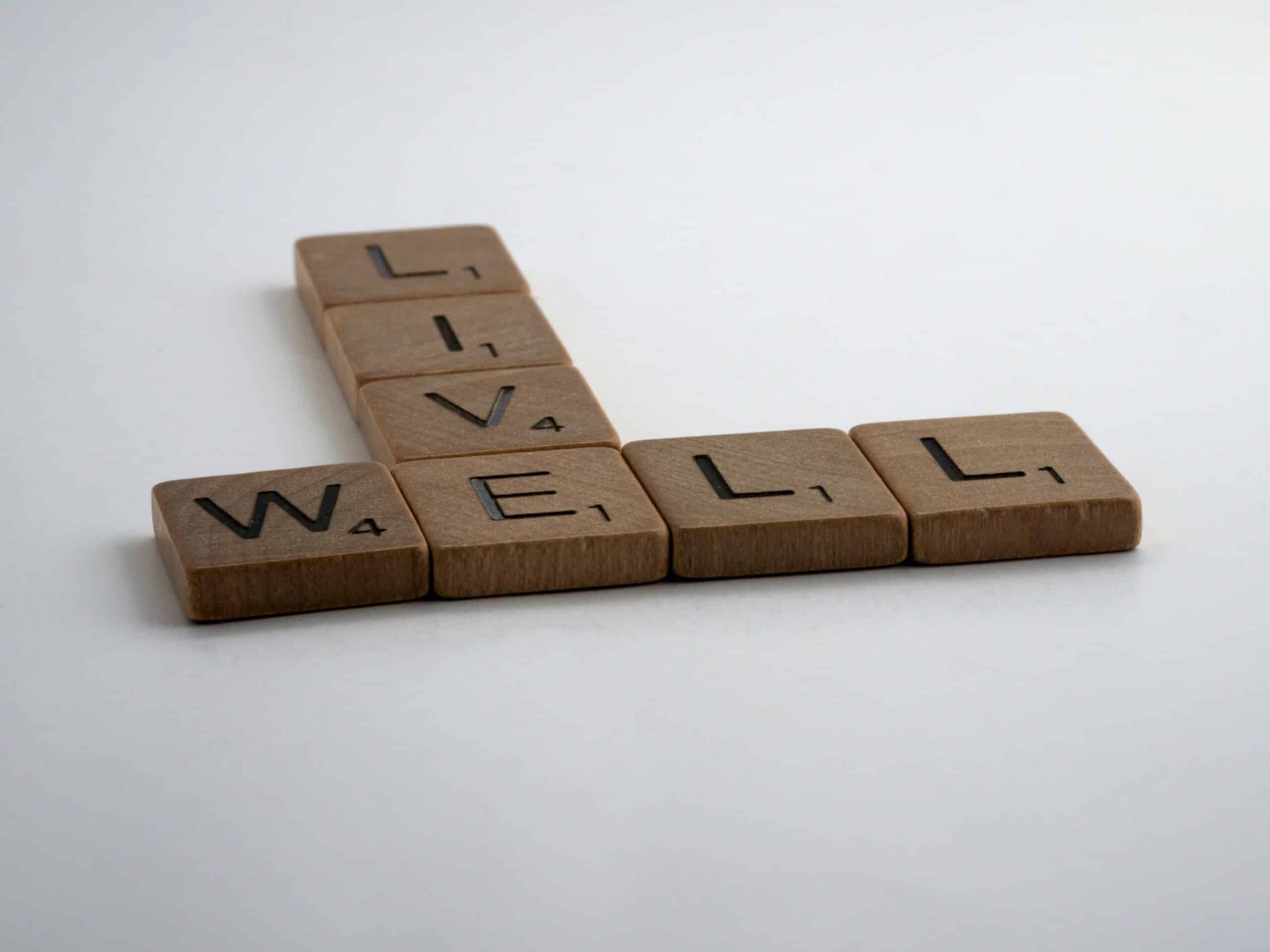 Four Scrabble tiles spelling "WELL" are arranged horizontally, with "LIVE" intersecting the "L," symbolising the importance of mental wellbeing. The tiles are set against a plain white background.