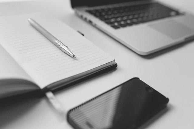 A silver pen rests on an open lined notebook beside a blurred laptop and smartphone on a white desk, evoking the convenient experience of choosing to buy medicine online from a trusted online chemist.