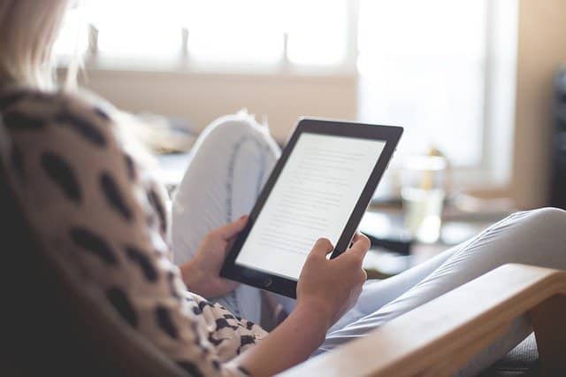 A person sits comfortably in a chair, holding a tablet and reading about teenager career options. The background is softly lit by natural light coming through a window. The person is wearing a patterned shirt and light trousers.