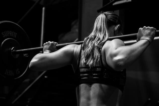 A woman with long hair and a sports bra lifts a barbell across her shoulders in a gym. The black-and-white image highlights her muscles and focused posture, reflecting the dedication often seen in those exploring CBD fitness for enhanced performance.