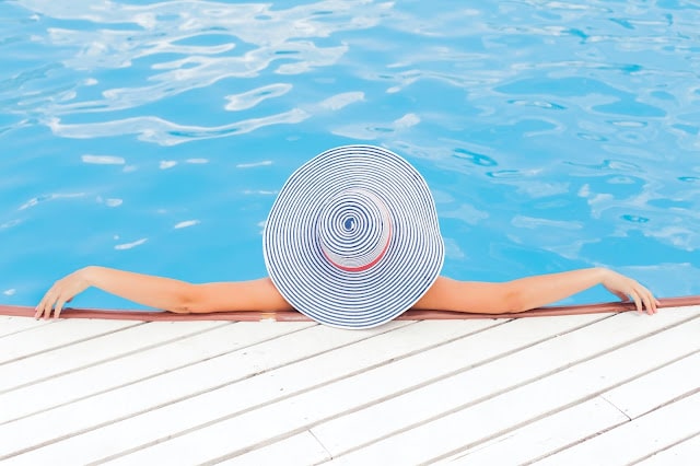 Person wearing a large, round striped sunhat relaxing in a swimming pool, arms stretched out along the pool edge, embracing the summer groove, with blue water and a white wooden decking visible.
