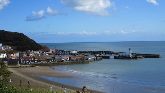 Coastal town with colourful buildings, a sandy beach perfect for outdoor fun, and a curved harbour wall with a white lighthouse. Calm blue sea and sky with scattered clouds; green hills rise on the left.