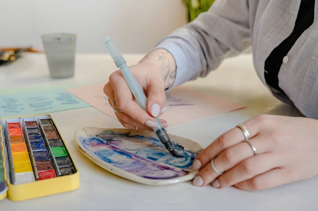 A person with rings and a tattoo uses a brush pen to mix blue and purple watercolour paint on a ceramic palette, preparing vibrant art for a summer party, with a watercolour set and paper on a white table nearby.
