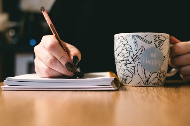 a woman writing in a notebook with a cup in her other hand