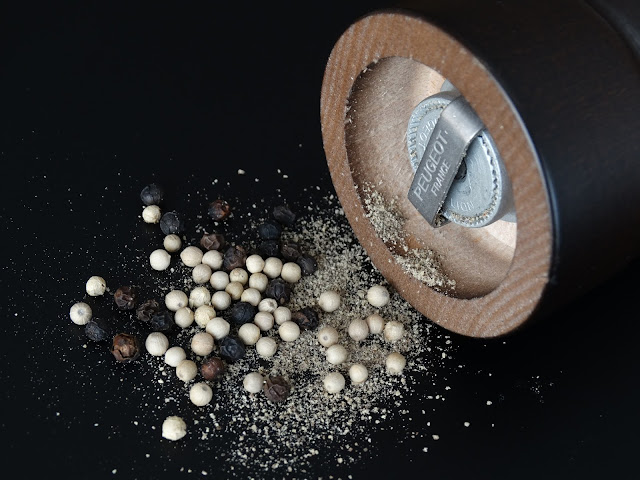 A close-up of a pepper mill on its side with mixed whole black and white peppercorns, some ground pepper spilt on a black surface—showcasing how useful pepper mills are for enhancing your spices.