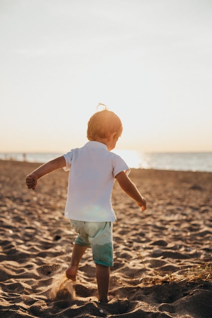 A young child, perfect for under 5's summer day trips, walks barefoot on a sandy beach towards the sea at sunset, with the sun low in the sky and soft light casting a warm glow.