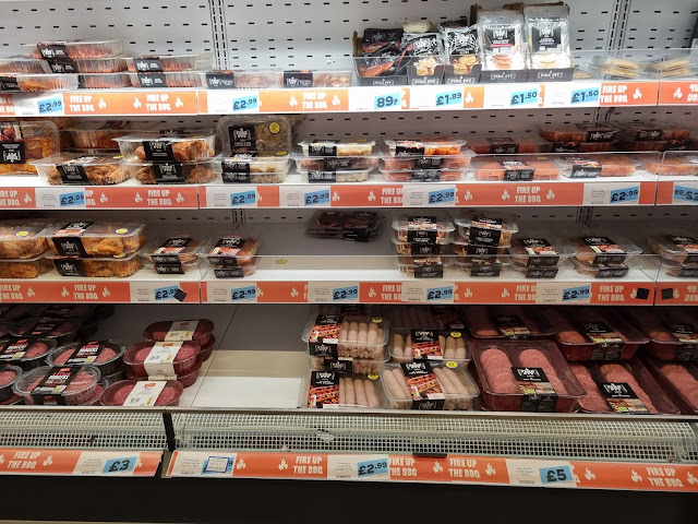 Shelves at Jack's Supermarket display various packaged meats—sausages, burgers, and chicken—all labelled with prices and "fire up the BBQ" promotional signage for the End of Term. The shelves are well-stocked and organised.