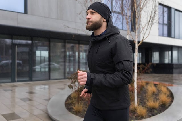 A man wearing a black jacket, hat, and trousers jogs on a paved pavement near a modern building with large windows, overcoming the elements as he passes through winter landscaping.