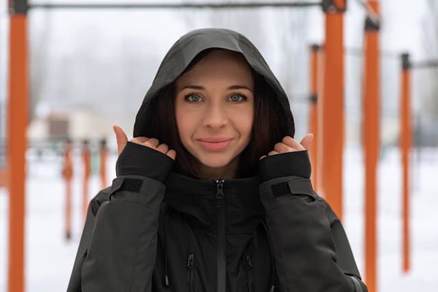 A woman with dark hair and blue eyes stands outdoors, facing the snowy background and orange bars. Wearing a black hooded jacket, she holds the edges of her hood with both hands, overcoming the elements with calm determination.
