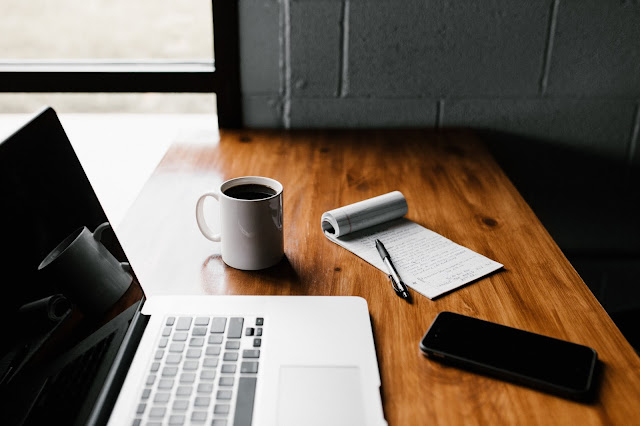 A laptop, mobile phone, white coffee cup and open notebook sat on a desk