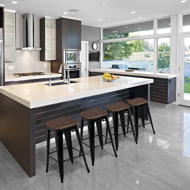 Modern kitchen with a large island, four safe bar stools, stainless steel appliances, sleek dark cupboards, and large windows letting in natural light. A bowl of fruit sits on the worktop.