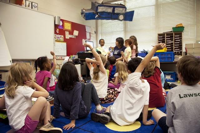A number of children sat on the floor around a teacher, some of the children have their hands up