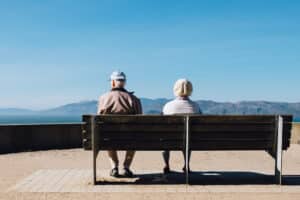Two elderly people sit on a bench facing the sea, with mountains in the distance under a clear blue sky—enjoying peaceful UK holidays perfect for senior relatives seeking relaxation outdoors.