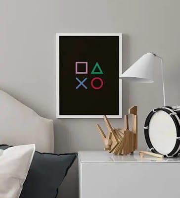 A bedside table with a drum, a modern wooden sculpture, and a white lamp sits beside a bed in this video game bedroom. Above, a framed print displays PlayStation controller symbols on a black background, adding themed bedroom flair.