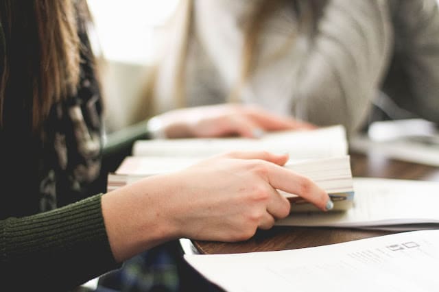 Close-up of a person’s hand holding an open book, with another reading nearby. Papers and books are spread out on the table, hinting at parental pointers for a child preparing for overseas studies during a focused work session.