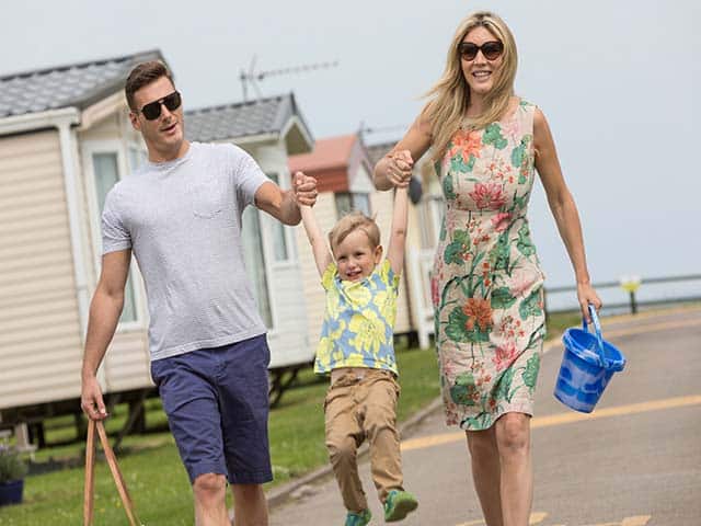 A smiling man and woman stroll outside by caravans at a Yorkshire Holiday Park, holding hands with a young boy and swinging him playfully. The woman carries a blue bucket, and they all look happy and relaxed.
