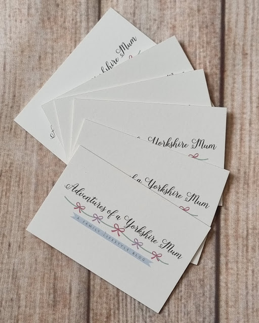 A stack of business cards reading "Adventures of a Yorkshire Mum" with a pink bow design, displayed on a wooden surface—perfect for anyone seeking an authentic Aura Print review.