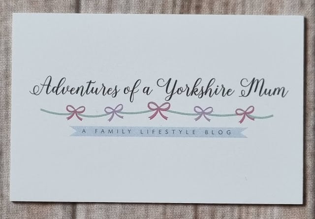 A white business card from Aura Print features "Adventures of a Yorkshire Mum" above pastel pink and purple bows on a green line, with "A Family Lifestyle Blog" in a blue banner. The card rests on a beige fabric background, perfect for review.