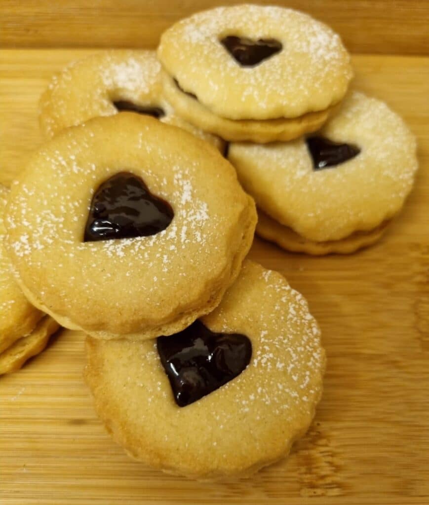 Round cherry jammy biscuits with heart-shaped cut-outs, filled with dark jam and dusted with icing sugar, are stacked on a wooden surface—perfect for Bank Holiday baking fun.