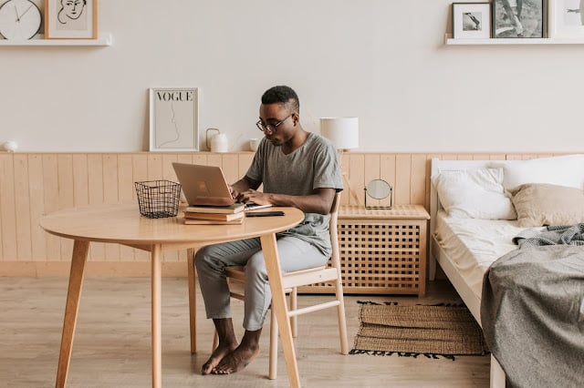 A man sat at a desk, working on a laptop, next to a bed