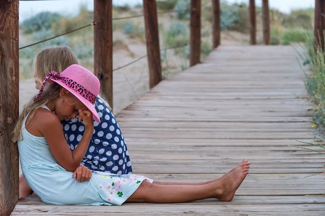 Two young girls sit on a wooden boardwalk. One in a light dress and pink hat rests her head on her hand, looking down—reminding us of the importance of support for children’s mental health in everyday moments.