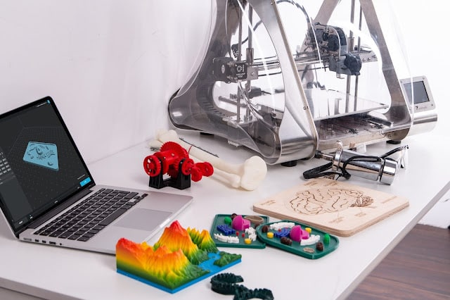 A 3D printer sits on a white desk beside a laptop displaying 3D modelling software. Colourful 3D-printed objects and robot parts, perfect for exploring STEM subjects, are scattered nearby along with a wooden board with engraved designs.