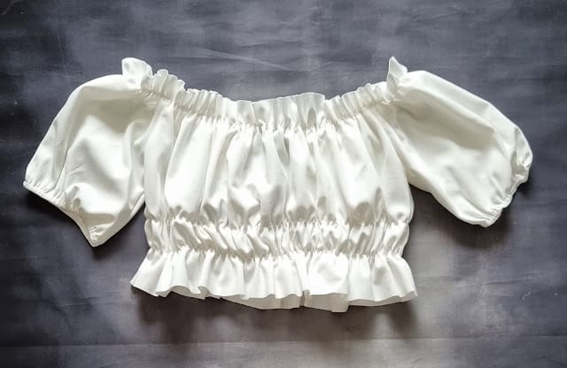 White off-the-shoulder crop top with short puffed sleeves and gathered, elasticated hems, laid flat on a dark grey background—a September fashion must-have and one of our Femmeluxe favourites.