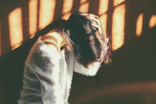A person with long hair sits in soft sunlight, head bowed and hand resting on their head, suggesting a moment seeking migraine relief. Shadows and light create patterns on the wall and their clothing, enhancing the contemplative mood.