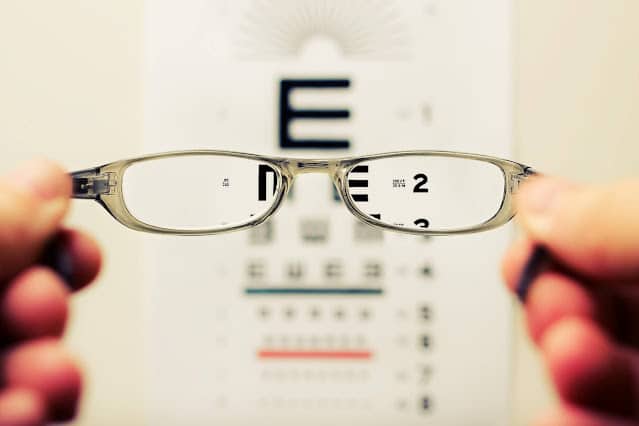 A pair of spectacles is held in front of an eye chart, bringing the chart into focus through the lenses while the rest of the image remains blurred—a clear reminder of the importance of regular eye care for maintaining optimal vision.