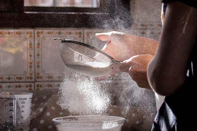 A person sifting Shipton Mill flour through a metal sieve into a bowl, with flour dust floating in the air and sunlight streaming in from a nearby window—a perfect moment of baking, framed by patterned kitchen tiles and a measuring jug.