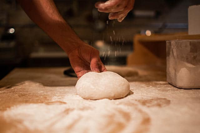 A person sprinkles Shipton Mill flour onto a round ball of dough on a wooden surface, preparing to bake. The scene is warmly lit, with flour scattered across the workspace—perfect inspiration for new recipes.